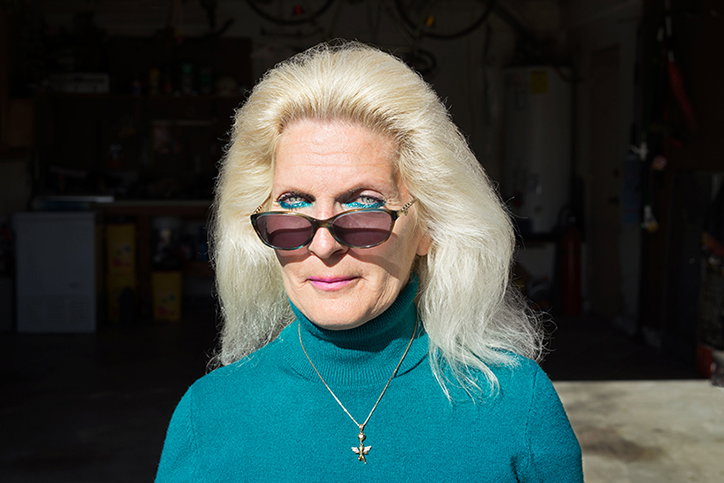 A person with voluminous blonde hair wears dark sunglasses and a teal turtleneck. They look directly at the camera against a blurred, dark indoor background.