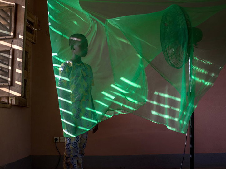A person stands behind a billowing sheet of translucent green fabric. A fan blows the material, creating ripples and casting striped shadows across the scene.