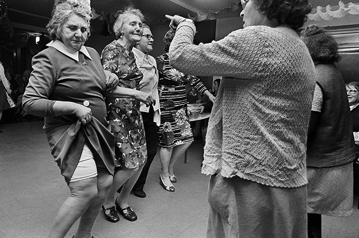 A group of people dance together in a brightly lit hall. In the foreground, a person in a grey dress lifts the hem of their skirt as they move.