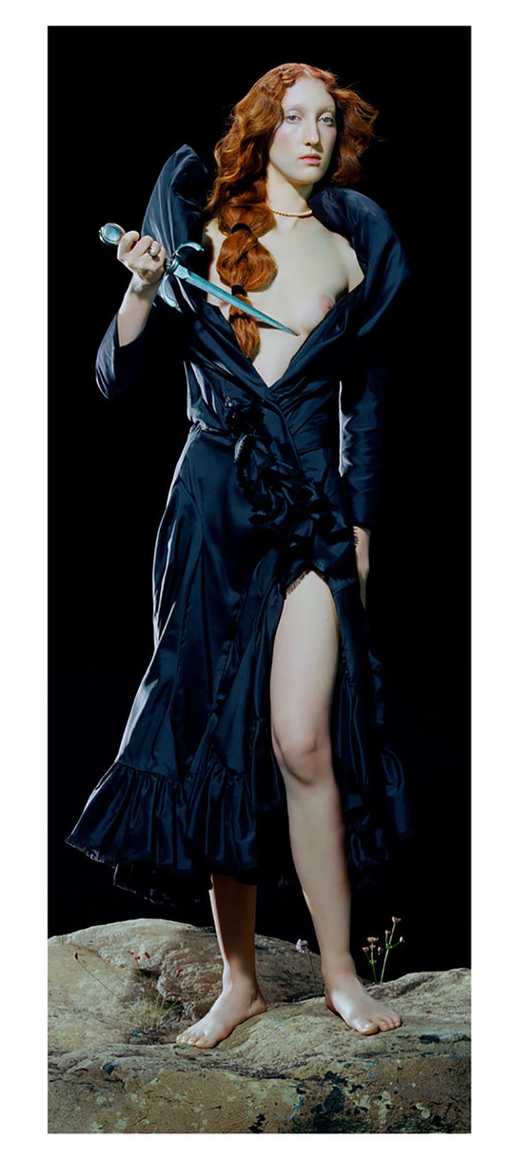 A person with long red hair stands barefoot on a rock against a black background. They wear a dark, flowing gown and hold a dagger across their chest.