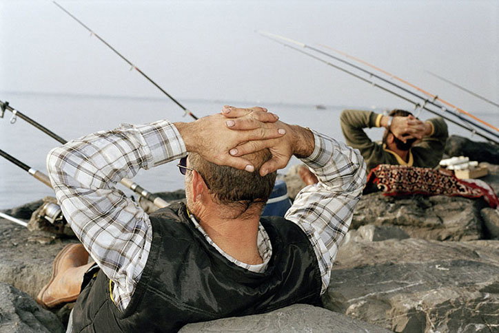 Two people lounge on a rocky shoreline, leaning back with their hands behind their heads. Several fishing rods lean against the rocks, pointing up towards a pale sky.