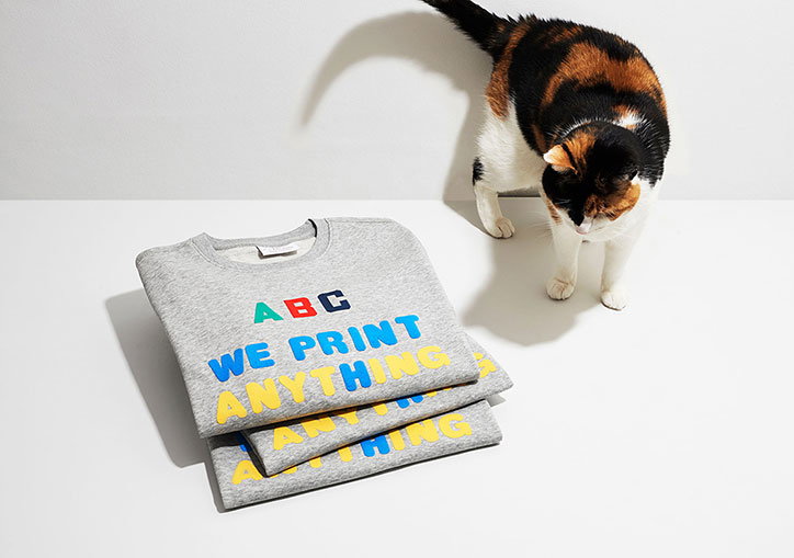 A calico cat looks down at a stack of grey sweatshirts. The top sweatshirt features colourful lettering that reads “ABC WE PRINT ANYTHING”.