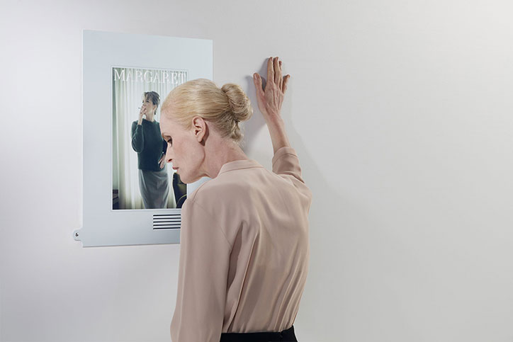 A person with blonde hair tied in a bun leans against a white wall, their hand pressed flat against the surface. Beside them hangs a portrait of a person with short hair, topped with the word “MARGARET”.