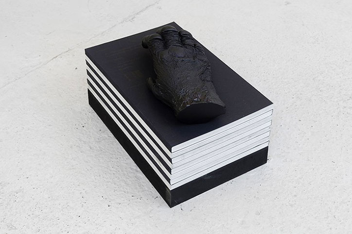 A sculpture of a dark, textured hand rests on top of a stack of books. The books have black covers and white pages, creating a striped effect against a plain white background.