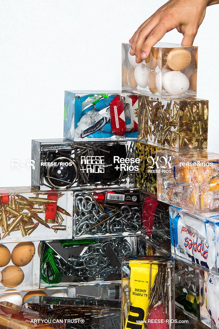 A hand reaches for a clear acrylic cube filled with white and brown eggs. The cube sits atop a precarious stack of similar boxes containing various objects, including bullets, nails, chains and tubes of adhesive. The boxes feature black labels with white text reading “Reese/Rios” and “Reese + Rios”. At the bottom of the image, a banner reads “ART YOU CAN TRUST®” and “www.reese-rios.com”.