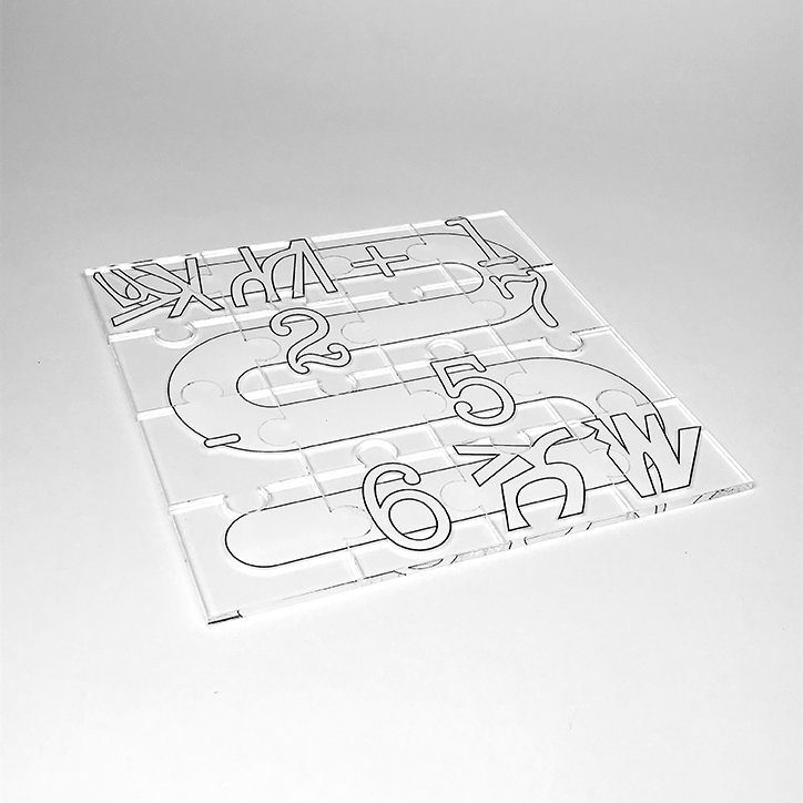 A clear acrylic slab etched with a winding path of lines and numbers. The path curves across the surface, featuring the numbers “1”, “2”, “5” and “6” alongside various abstract symbols.