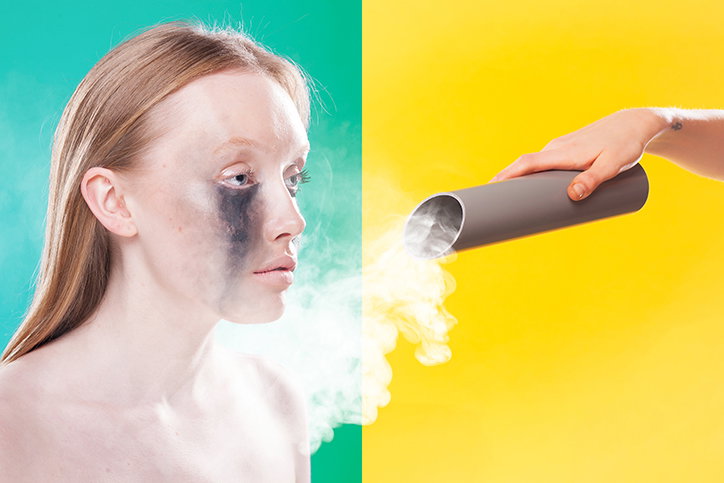 A person with black makeup smeared around one eye looks towards a grey tube. A hand holds the tube, from which a cloud of white smoke blows across the person’s face. The background is split vertically between green and yellow.