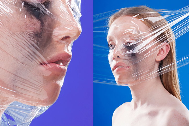 Two portraits of a person with clear plastic wrap stretched across their face. They wear heavy black makeup around their eyes against a bright blue background.