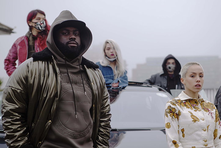 Five people stand together outdoors against a grey, misty backdrop. In the foreground, a person in a hooded olive jacket looks directly at the camera, while another person in a white floral jacket stands beside them.