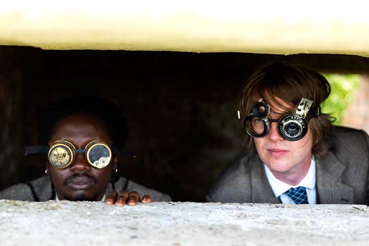 Two people peer out from a narrow concrete opening, both wearing oversized, experimental eyewear. The person on the left wears round glasses with metallic discs, while the person on the right wears a complex device resembling camera lenses.