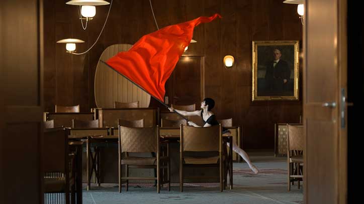 A person in a black dress waves a large red flag in a wood-panelled boardroom. They stand among a series of empty chairs and tables, with a framed portrait hanging on the wall behind them.