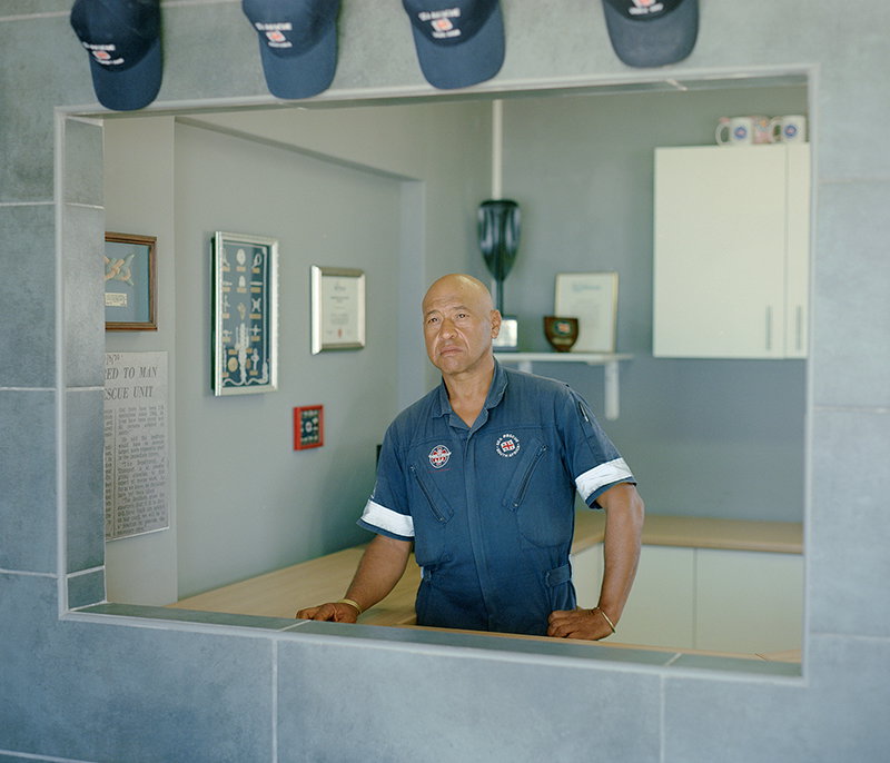 A person in a navy blue uniform stands behind a service counter, looking directly at the camera. They are framed by a rectangular opening in a grey tiled wall, with framed certificates and navy caps visible in the background.