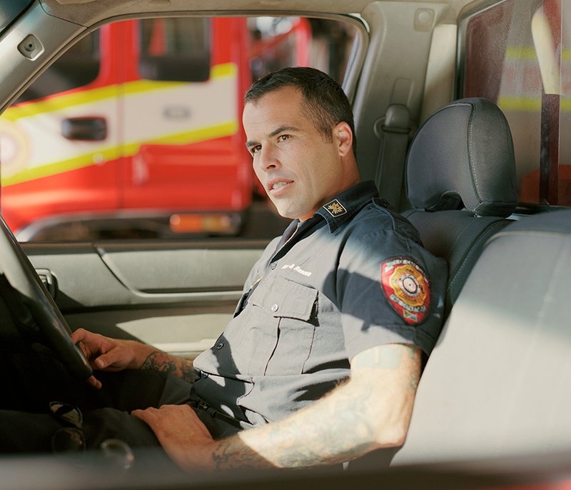A person in a dark grey firefighter's uniform sits in the driver's seat of a vehicle. Through the window, a red fire engine is visible in the background.