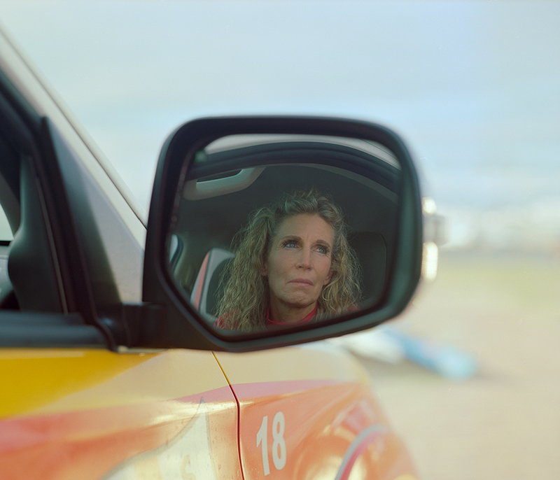 A person with long curly hair is reflected in a car wing mirror, looking ahead with a serious expression. The yellow car body below the mirror is marked with the number “18”.