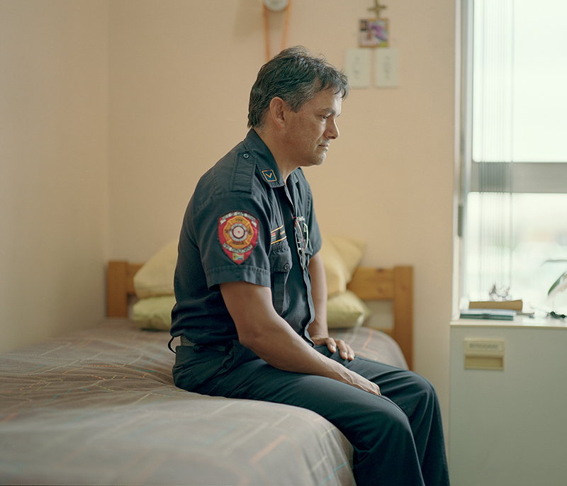 A person in a dark firefighter’s uniform sits on the edge of a bed, looking down and away from the camera. They are in a sparsely furnished room with a window to their right.