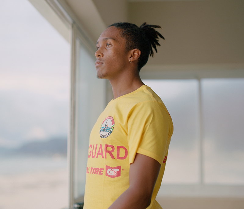 A person with dreadlocks stands in profile, looking out of a window. They wear a yellow T-shirt with a circular logo and the word “GUARD” printed in red.