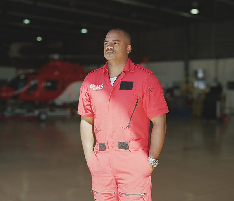 A person in a red flight suit stands with their hands in their pockets, looking off-camera. They wear a suit branded with the “Gams” logo, set against a blurred hangar with a red helicopter in the background.