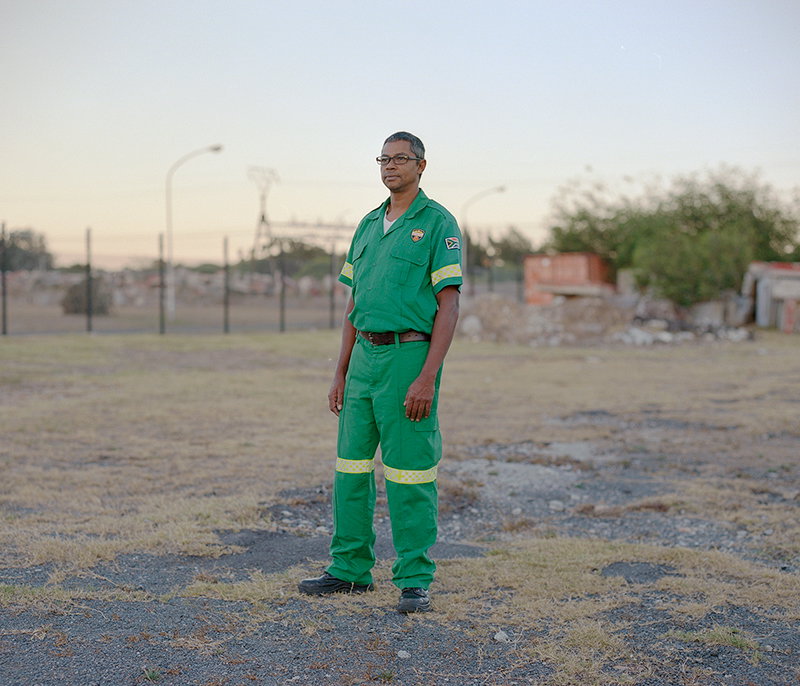 A person wearing a green utility jumpsuit and glasses stands in a wide, open gravel lot. In the background, a low fence and distant buildings sit under a pale sky.