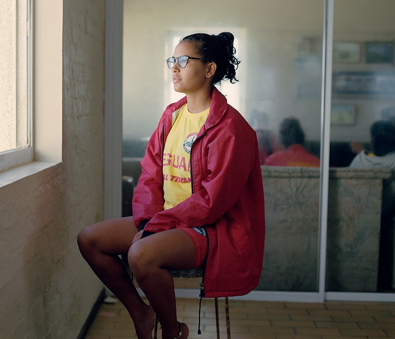 A person wearing glasses and a red jacket sits on a stool, looking out of a window. They wear a yellow T-shirt with the word “GUAN” visible on the front.