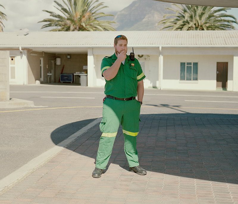 A person in a green utility uniform stands in a paved courtyard, speaking into a handheld radio. Behind them are low white buildings, palm trees and a distant mountain.