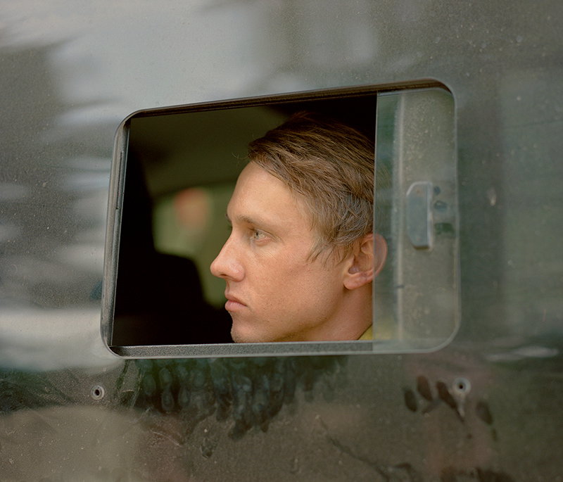 A person is seen in profile, looking out through a small, rectangular window in a dark grey wall. Their expression is neutral and focused.