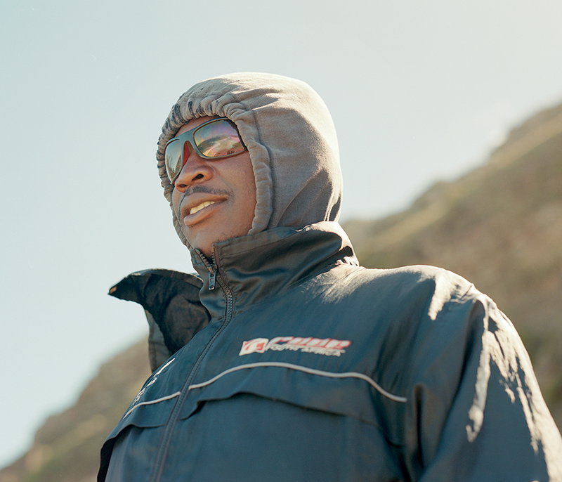 A person wears a grey hood and sunglasses, looking off to the side. They are dressed in a black technical jacket with a small red and white logo on the chest.