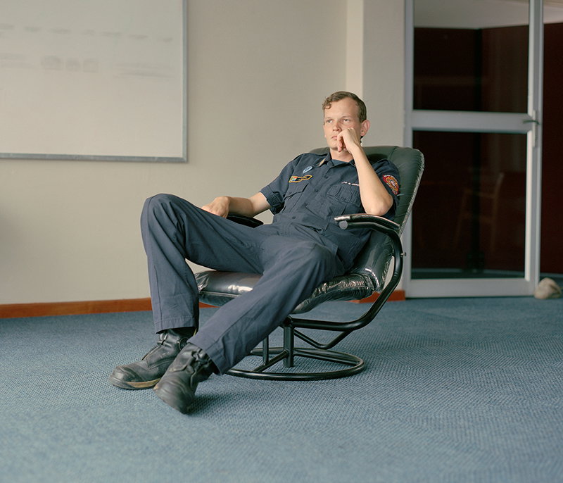 A person in a dark blue uniform sits reclined in a black leather lounge chair. They lean their head on one hand, looking thoughtfully off to the side.