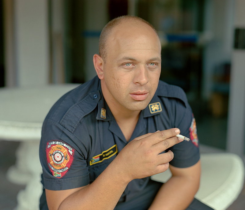 A person in a dark blue uniform sits and looks towards the camera while holding a cigarette. A patch on their sleeve reads “City of Cape Town Fire & Rescue Department”.