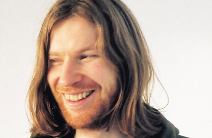 A person with long hair and a beard smiles, looking away from the camera. They are set against a plain white background.