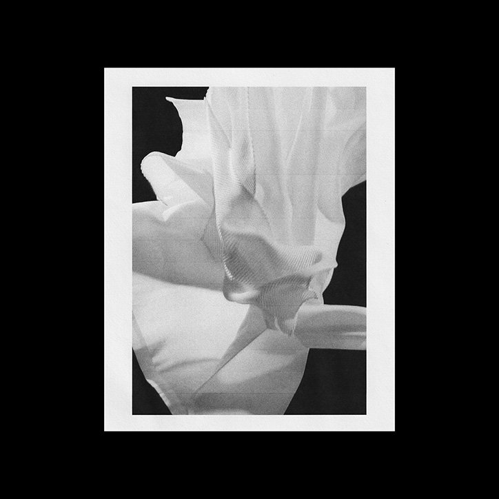A black and white photograph of draped white fabric, folded and twisted into abstract shapes. The material is set against a stark black background and framed by a wide white border.