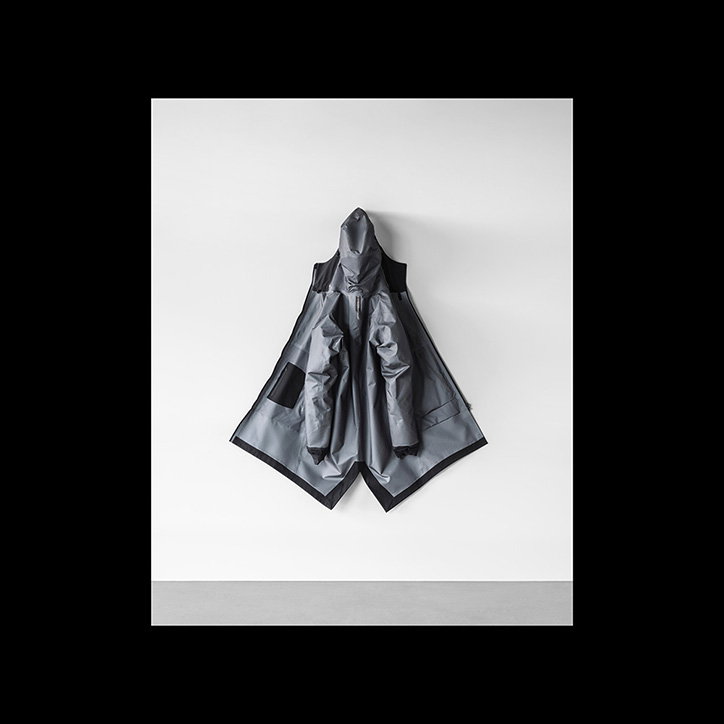 A grey, hooded technical jacket is displayed flat against a plain white wall. The garment has a structured, angular silhouette with wide panels and a high collar.