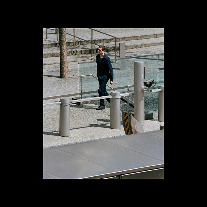 A person in a dark navy suit walks across a grey concrete plaza. They are positioned between several silver metal bollards and a glass railing.