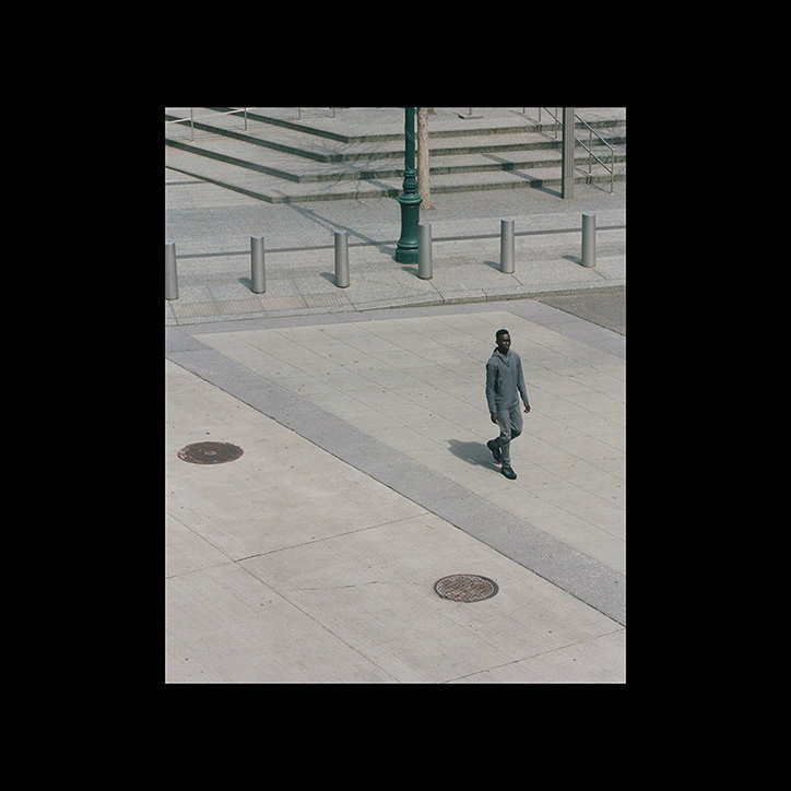 A person in a grey suit walks across a wide, pale concrete plaza. In the background, a set of stone steps is lined with short metal bollards and a single green lamp post.