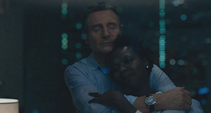 Two people embrace in a dimly lit room. Behind them, a window looks out onto a city skyline with blurred green and white lights.
