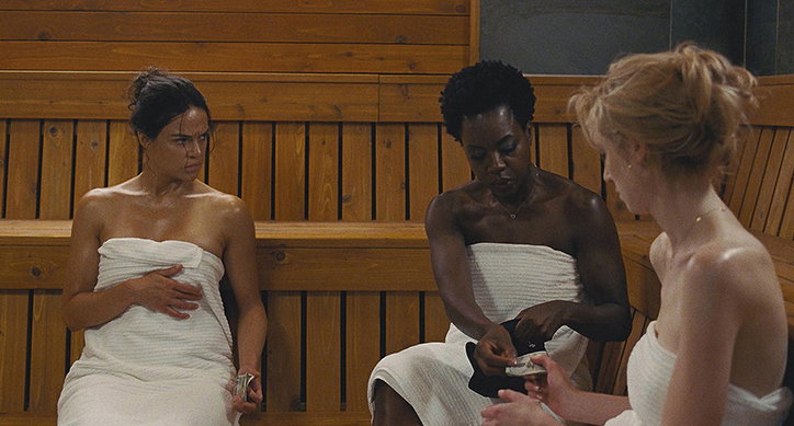 Three people wrapped in white towels sit on the wooden benches of a sauna. One person holds a small object while the others look on.