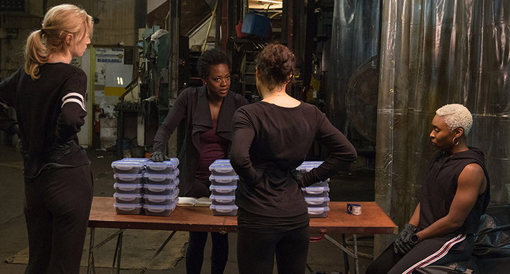 Four people gather around a wooden table in a dimly lit industrial space. Stacks of plastic food containers sit on the table as the group talks.