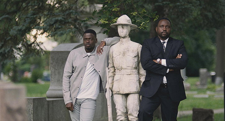 Two people stand on either side of a weathered stone statue of a soldier in a cemetery. One person leans against the statue in a light grey jacket, while the other stands with arms crossed in a dark suit.