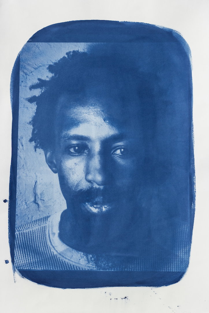 A cyanotype portrait of a person with curly hair and a beard, looking directly at the camera. The deep blue image is framed by a rough, hand-painted border on white paper.