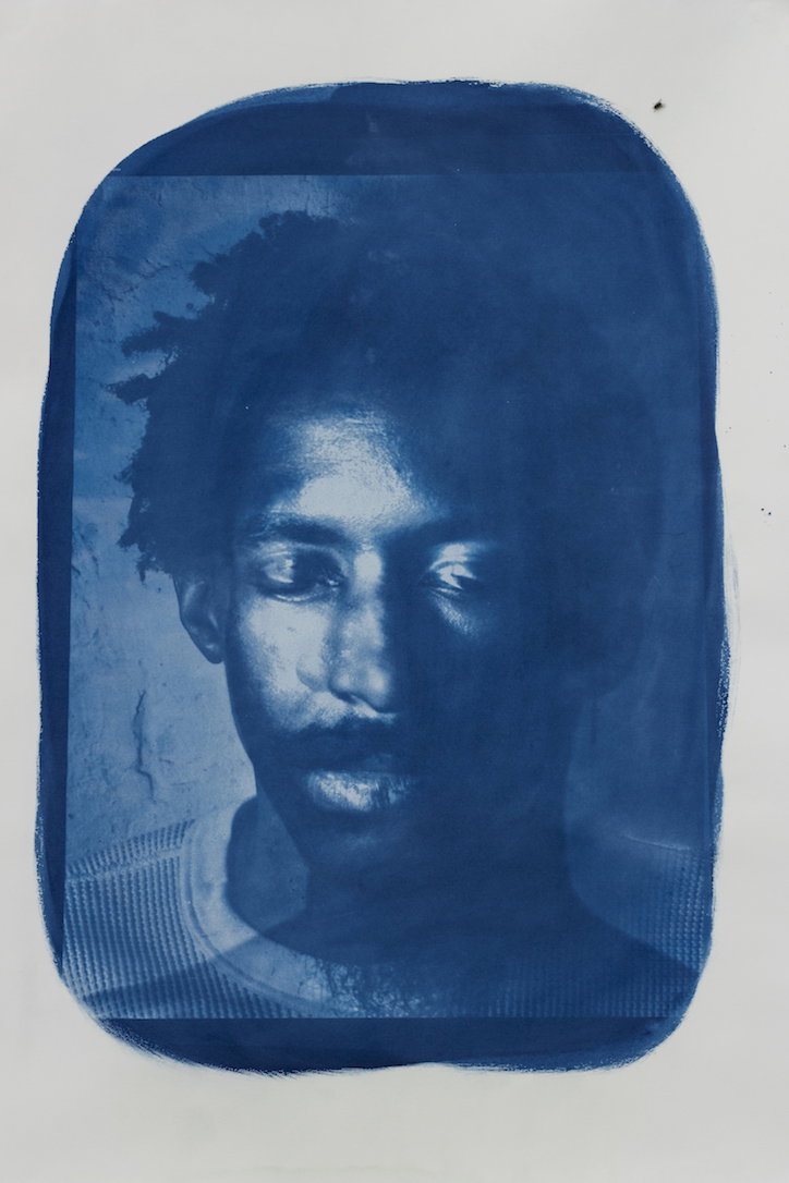 A cyanotype portrait of a person looking directly at the camera. The image is rendered in deep blue tones and framed by a rough, hand-painted border.