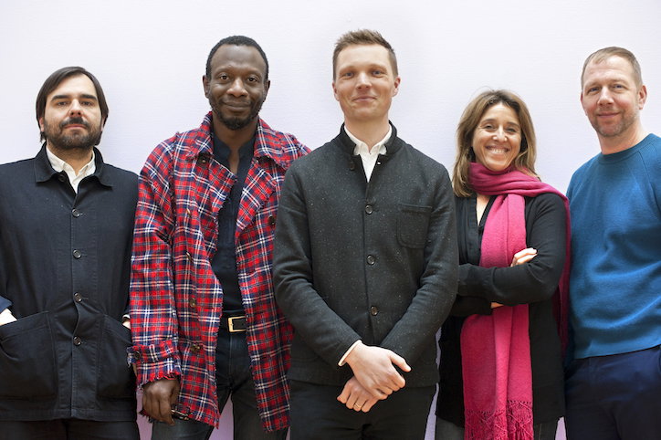Five people stand side by side against a plain white background. They wear a mix of casual and smart clothing, including a red plaid jacket and a bright pink scarf.
