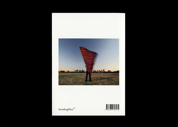 A book cover featuring a photograph of a person standing in a grassy field, holding a large, red fabric that billows upwards. A city skyline is visible on the horizon under a clear blue sky. The Sternberg Press logo and a barcode appear at the bottom.