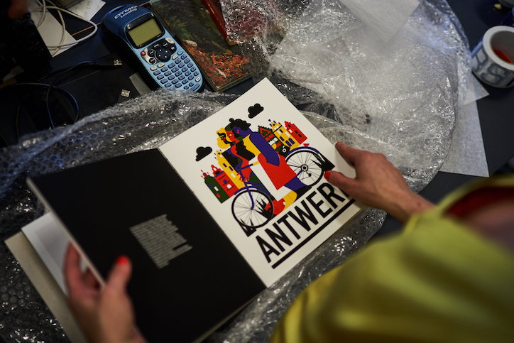 A person holds open a book to a page featuring a colourful illustration of a figure on a bicycle. Bold text below the image reads “ANTWERP”.