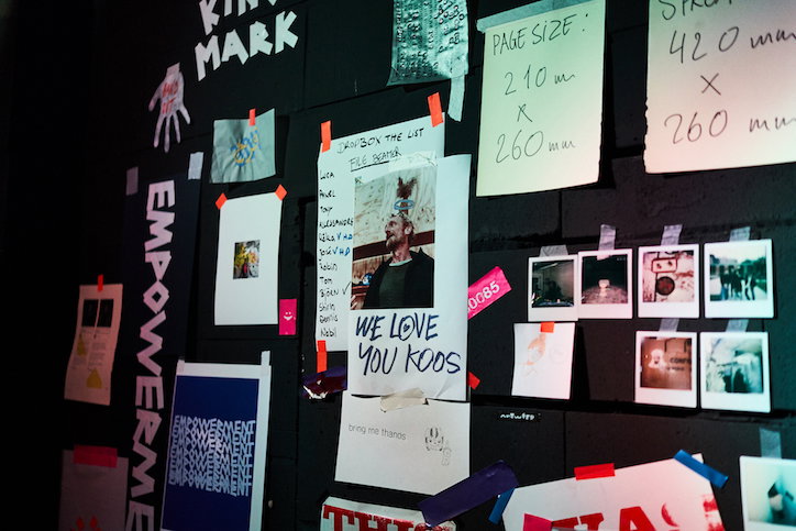 A black wall is covered in a collage of posters, notes and photographs. One central piece features a portrait of a person and the handwritten text “WE LOVE YOU KOOS”.