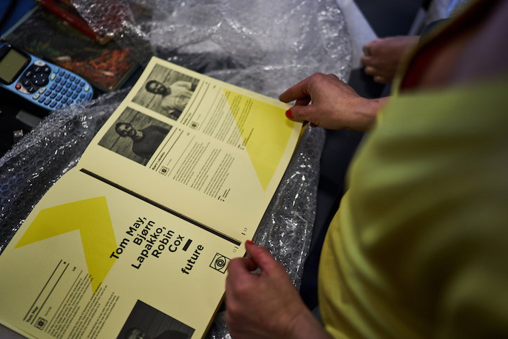 A person holds an open booklet with yellow and white pages over a sheet of bubble wrap. The page features black and white portraits and a list of names including “Tom May, Bjørn Lapakko, Robin Cox”.