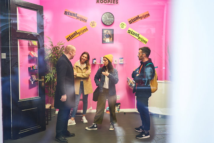 Four people stand in a room with bright pink walls, talking in a small group. Above them, a clock is surrounded by yellow banners that read “stuntprijzen”, with a sign above reading “koopjes”.