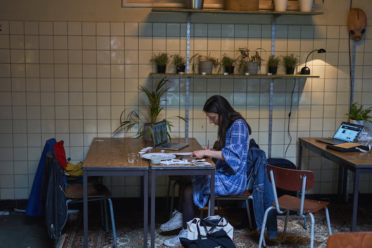 A person in a blue checked dress sits at a wooden table, working with papers and a laptop. They are in a room with white tiled walls and shelves holding several small potted plants.