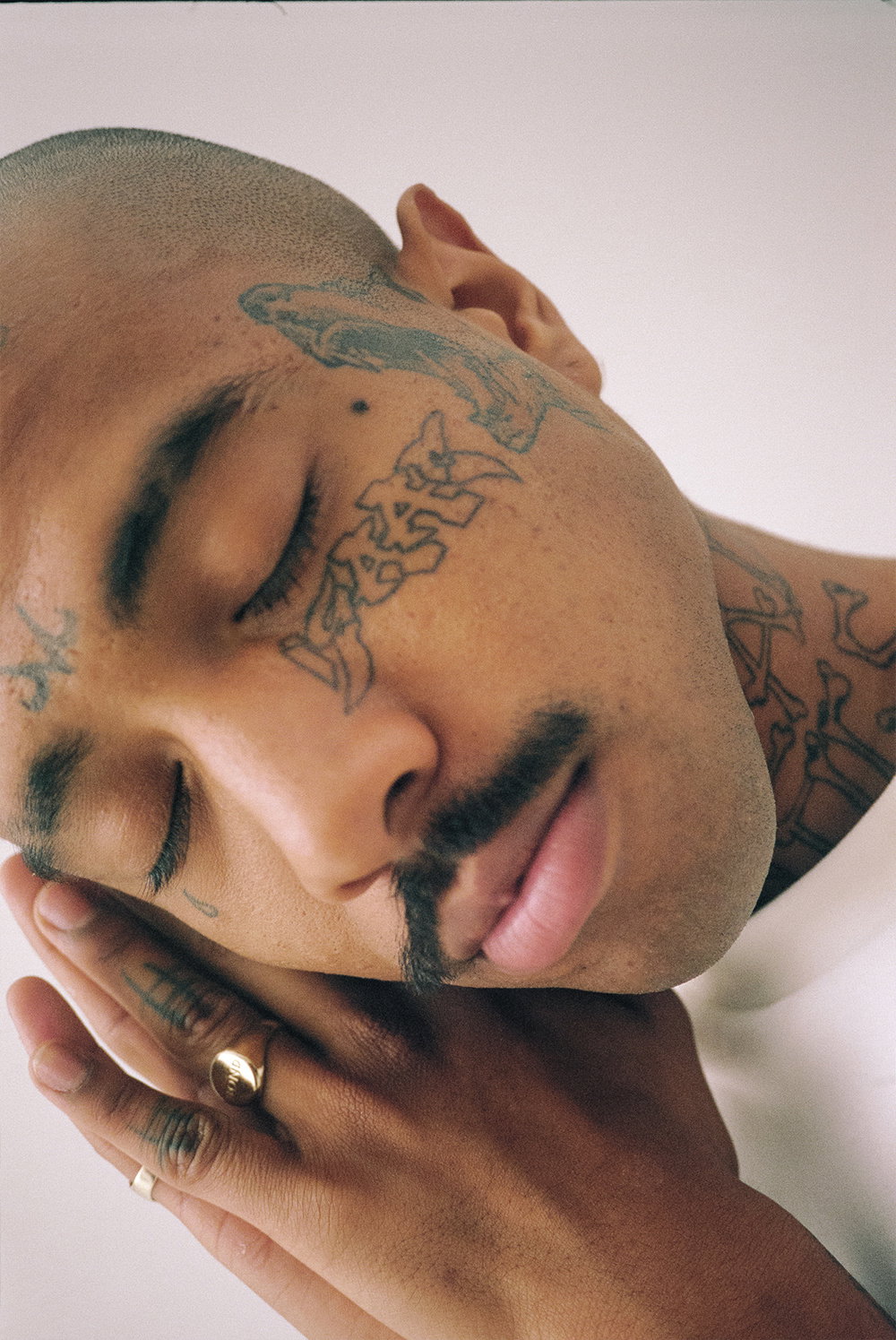 A close-up portrait of a person with their eyes closed and head tilted. They have extensive tattoos across their face, neck and hands, and wear a gold ring.