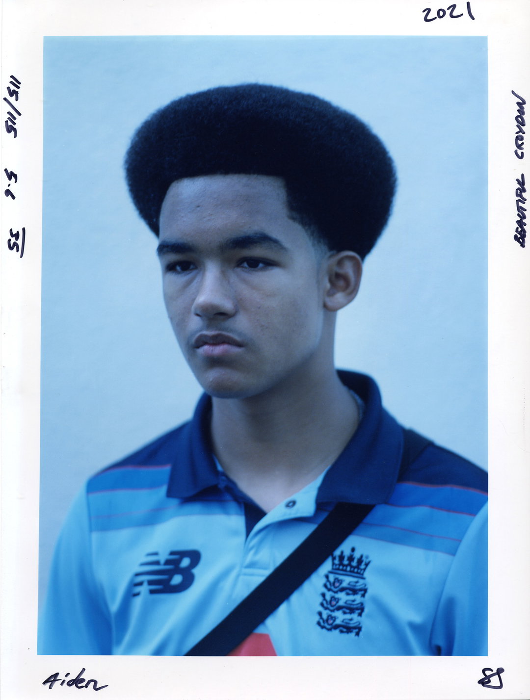A person with a high-top fade haircut looks off-camera with a neutral expression. They wear a light blue New Balance sports shirt with a dark blue collar.