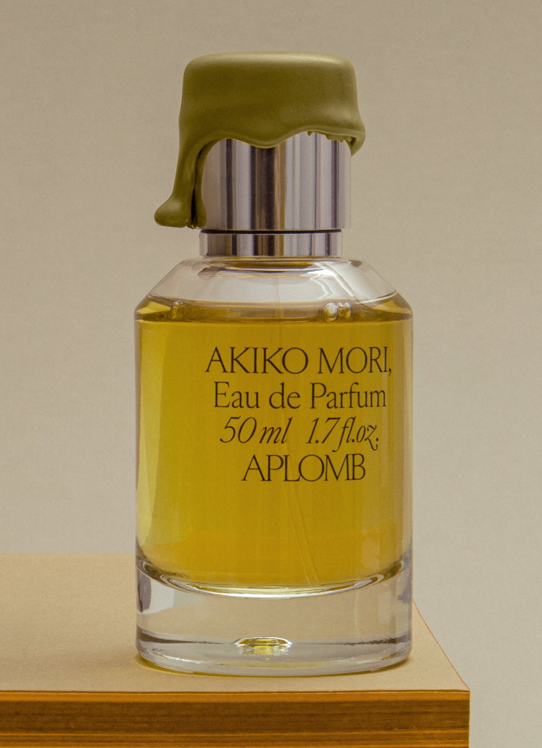 A glass bottle of yellow perfume sits on a tan surface. The label reads “AKIKO MORI, Eau de Parfum 50 ml 1.7 fl.oz. APLOMB”. A gold-coloured cap is sculpted to look like melting wax dripping down the silver neck.