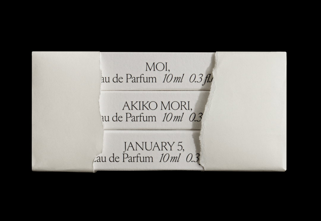Three white paper strips are layered horizontally against a black background. Each strip features serif text reading “eau de Parfum 10ml 0.3 fl.” alongside the names “Moi,” “Akiko Mori,” and the date “January 5,”.